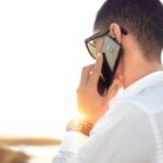 Man outdoors in Morocco talking on smartphone, wearing sunglasses and white shirt, backlit by sunset.